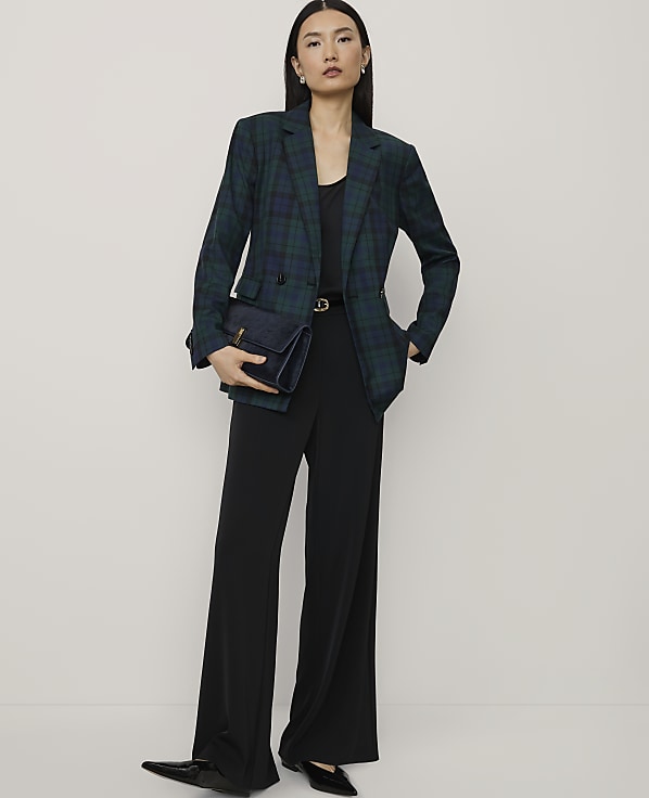 A woman wearing a green plaid blazer and black wide-leg trousers stands against a plain, light-colored background.