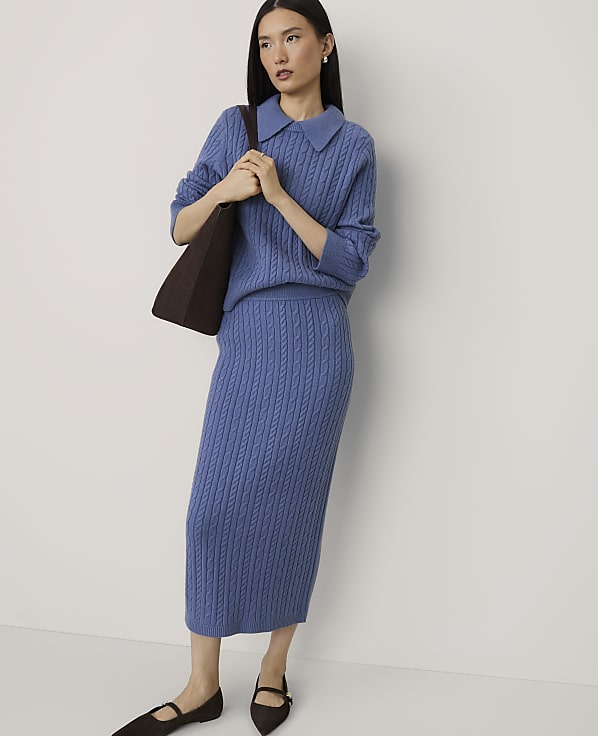 A woman wearing a blue cable-knit sweater and matching skirt stands against a plain white background.
