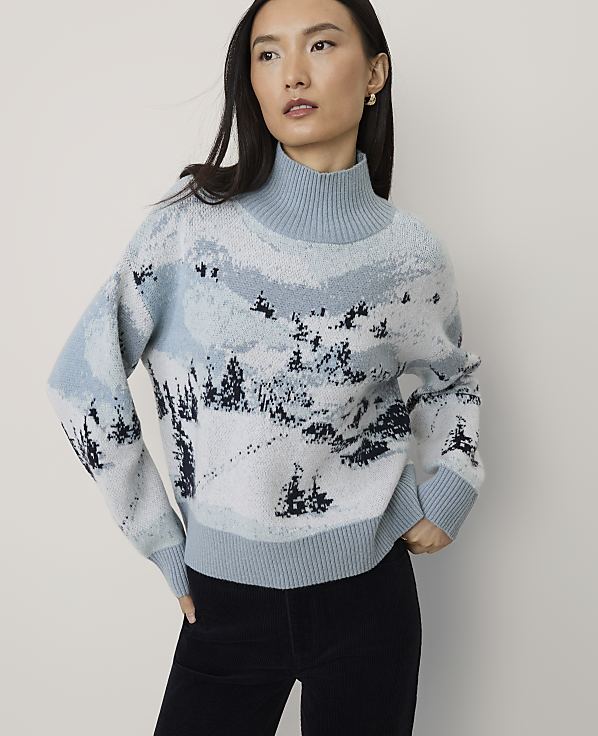 A woman wearing a light blue winter landscape patterned sweater and black pants stands against a plain white background.