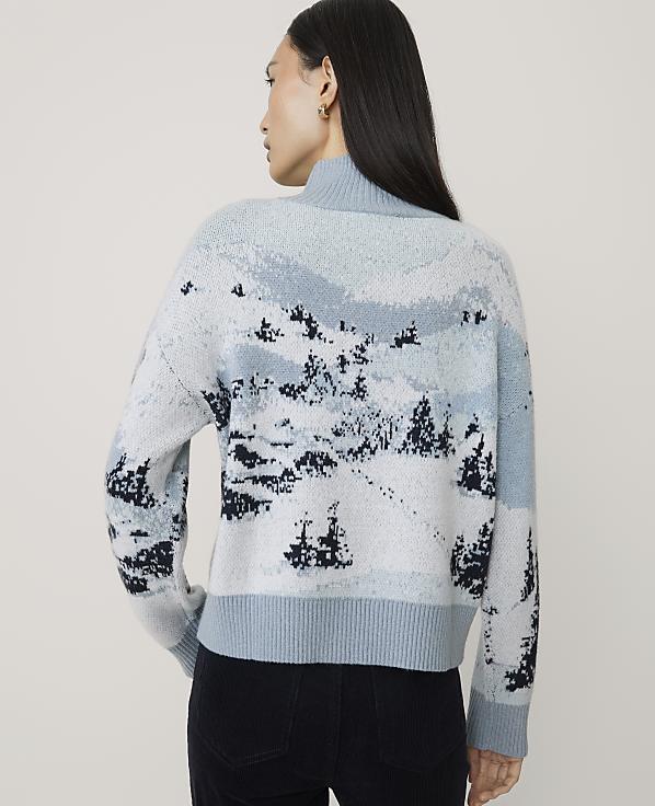 A woman wearing a light blue landscape-patterned sweater and black pants stands against a plain, light-colored background.