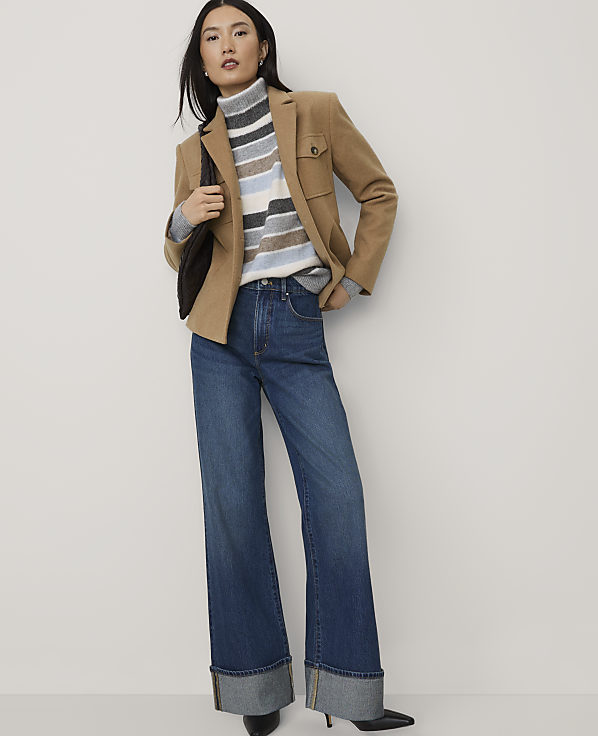 A woman wearing a tan blazer, a striped turtleneck, and wide-leg jeans stands against a plain white background.