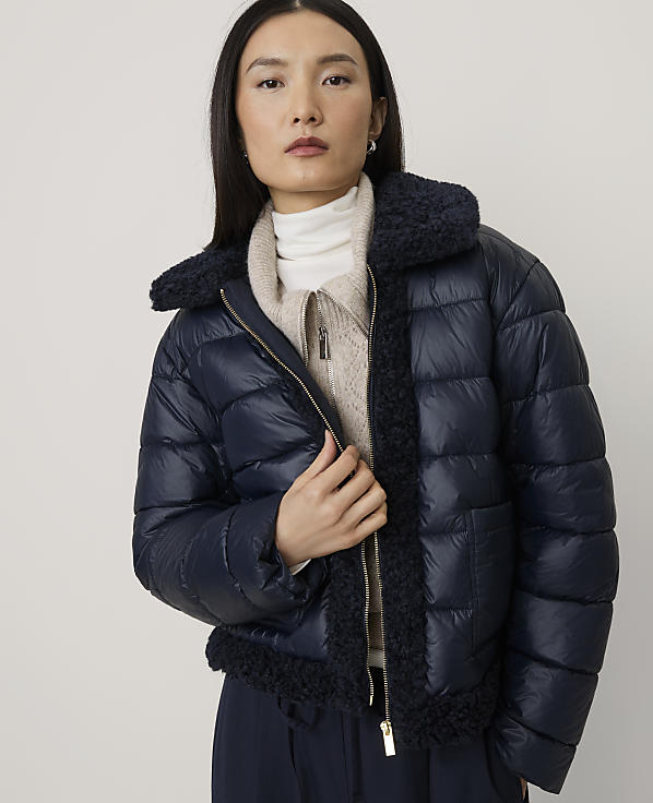 A woman wearing a navy blue puffer jacket over a beige sweater stands against a plain, light-colored background.