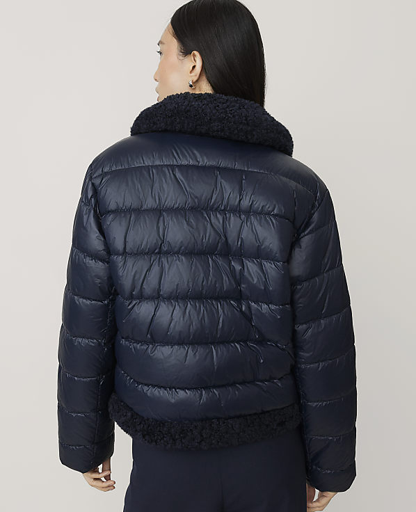A woman wearing a navy blue puffer jacket with a faux-fur collar stands against a plain, light-colored background.
