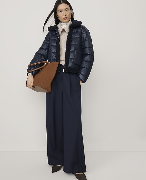 A woman wearing a navy puffer jacket and wide-leg trousers holds a brown handbag against a plain white background.