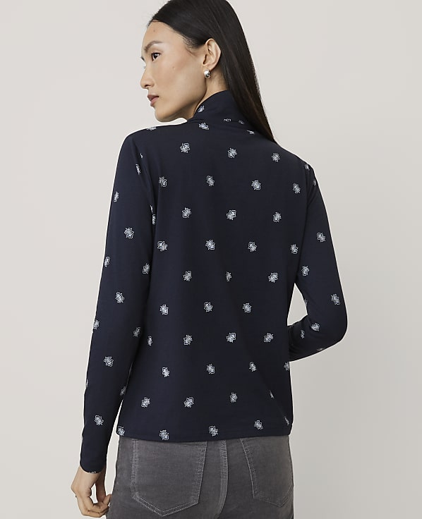A woman wearing a navy blue patterned long-sleeve shirt and grey pants stands against a plain light grey background.