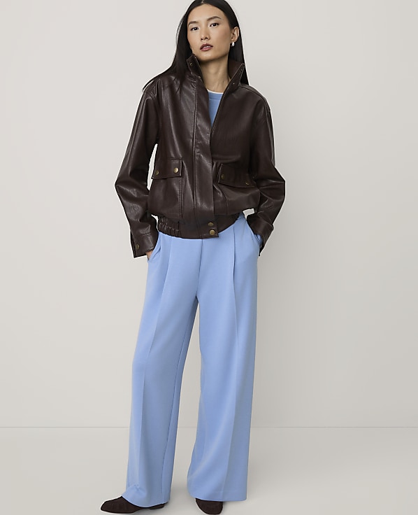 A woman wearing a brown leather jacket and wide-leg light blue trousers stands against a plain, neutral-colored background.