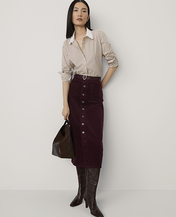 A woman wearing a checkered shirt, a burgundy corduroy skirt, and brown boots stands against a plain white background.