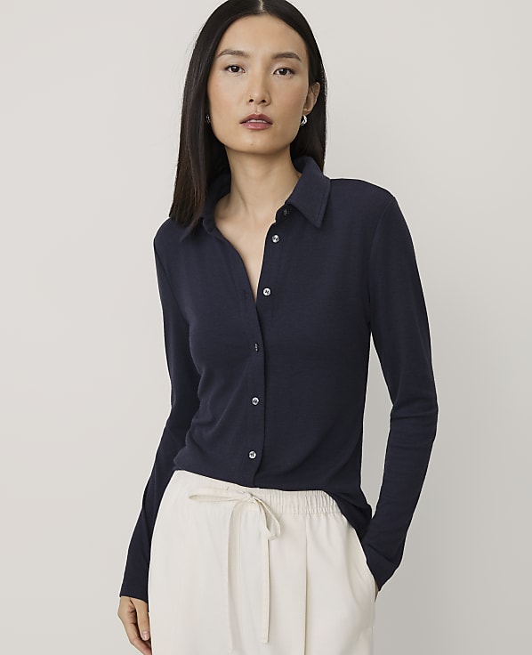 A woman wearing a navy blue long-sleeved collared shirt and white trousers stands against a plain light gray background.