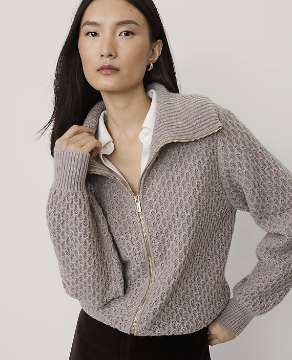 A woman wearing a textured mauve zip-up sweater over a white shirt stands against a plain light gray background.