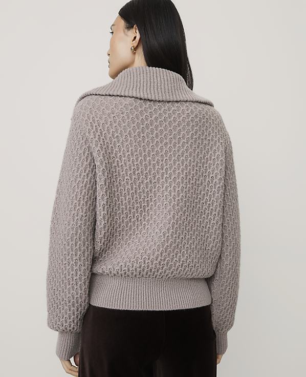 A woman wearing a textured taupe sweater and dark brown trousers stands against a plain light gray background.