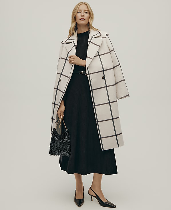 A blonde woman wearing a black pleated dress and a white windowpane check coat stands against a plain light grey background.