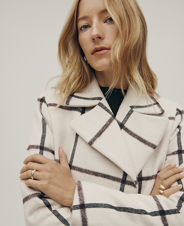 A blonde woman wearing a white and black checkered coat stands with her arms crossed against a plain, light-colored background.