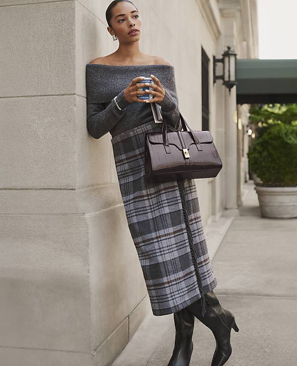 A woman in a grey off-the-shoulder top and plaid skirt leans against a stone wall on a city sidewalk.