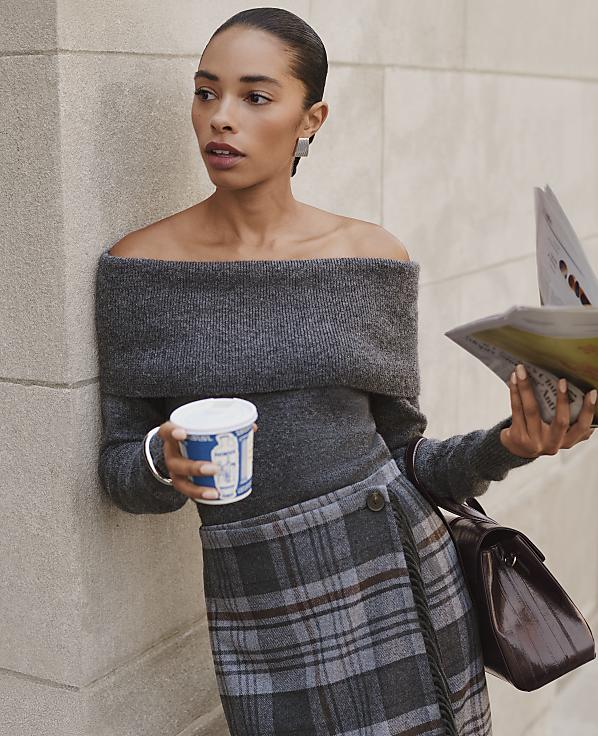 A woman in an off-the-shoulder grey sweater and plaid skirt holds a coffee cup and a newspaper while leaning against a light-colored stone wall.