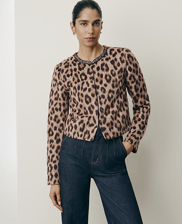 A woman wearing a leopard-print jacket and dark blue jeans stands against a plain, light-colored wall.