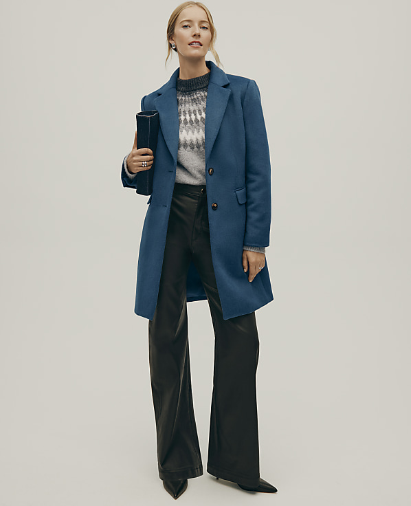 A woman wearing a blue coat, a grey patterned sweater, and black leather trousers stands against a plain, light-colored background.