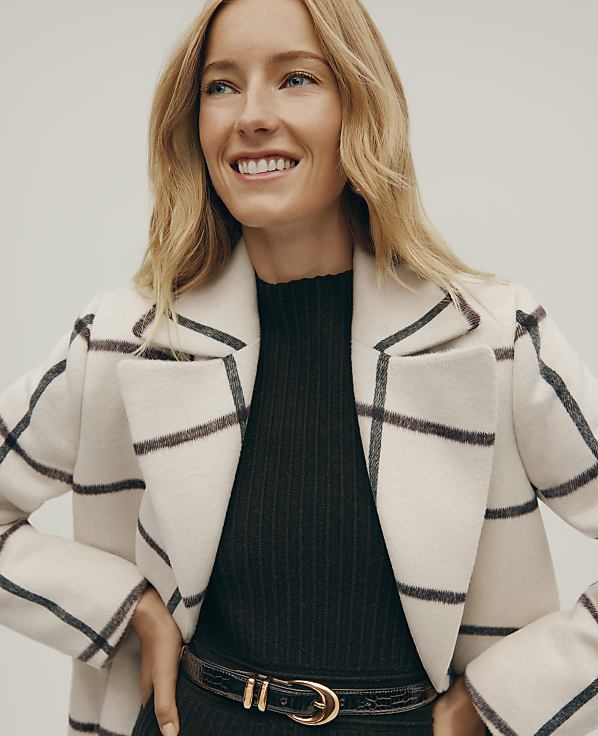 A smiling blonde woman wears a white grid-patterned coat over a black ribbed top and belt against a plain light gray background.