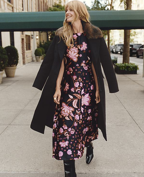A smiling woman wearing a floral dress and a black coat walks down a city sidewalk lined with buildings and trees.