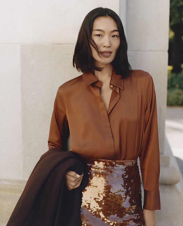 A woman wearing a brown satin shirt and a sequined skirt stands against a neutral-toned stone wall.