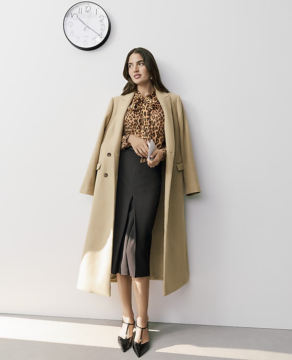 A woman wearing a leopard-print blouse, black skirt, and tan coat stands against a plain white wall with a clock hanging above her.