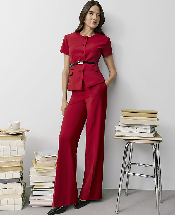 A woman in a red short-sleeved suit stands against a plain white wall, flanked by stacks of books on the floor and a metal stool.