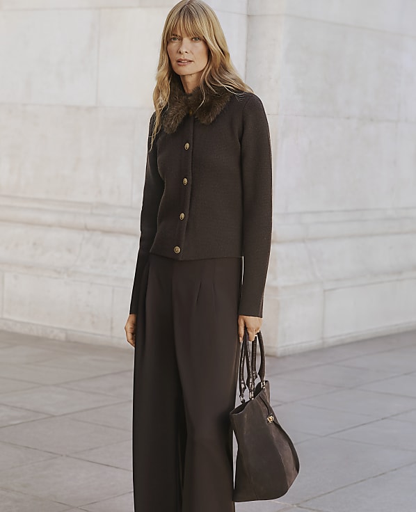 A woman wearing a brown button-up cardigan with a fur collar and wide-leg trousers stands holding a handbag against a light-colored stone wall.