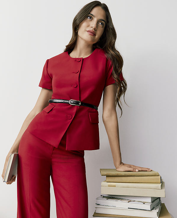 A woman in a red short-sleeved suit stands against a plain white background, leaning her hand on a stack of books.
