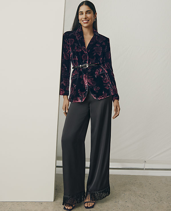A woman wearing a floral velvet blazer and wide-leg black trousers stands against a plain white wall and backdrop.