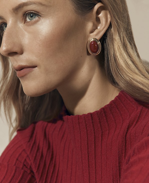 A woman wearing a red ribbed sweater and a red gemstone earring is positioned against a neutral, light-colored background.