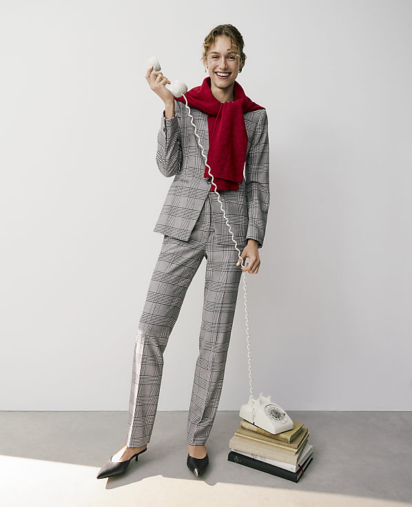 A smiling woman in a plaid suit and red scarf talks on a vintage white telephone resting on a stack of books against a plain white background.