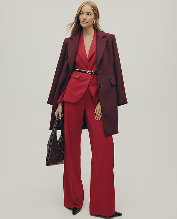 A woman wearing a red suit and a burgundy coat stands against a plain, light-colored background.