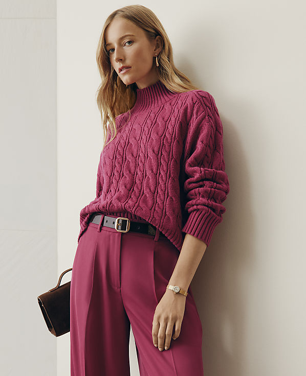 A woman wearing a magenta cable-knit sweater and matching trousers leans against a plain white wall.