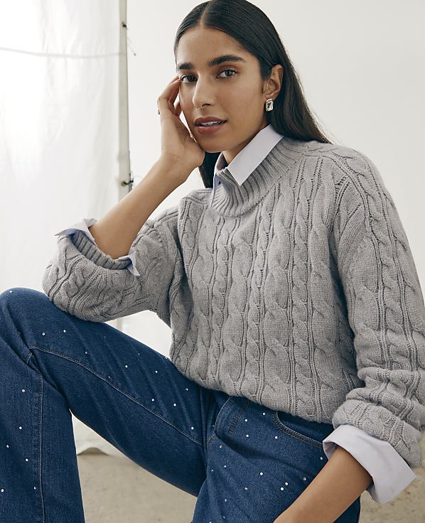 A woman wearing a grey cable-knit sweater over a white collared shirt and rhinestone-embellished jeans poses against a plain white backdrop.