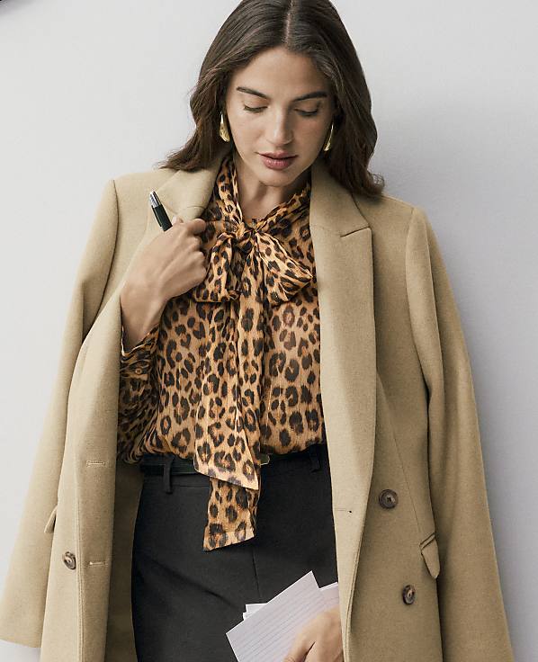 A woman wearing a leopard-print pussy-bow blouse, black trousers, and a tan coat holds a pen and papers against a plain light gray background.
