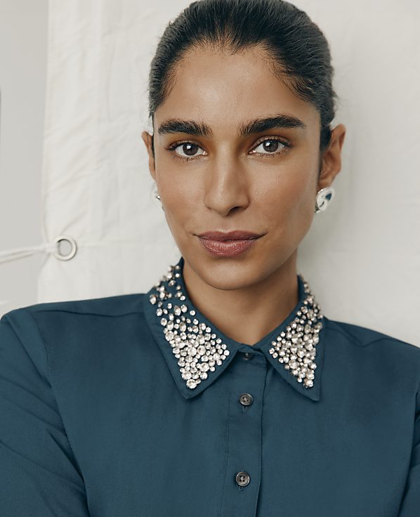 A woman wearing a teal shirt with a crystal-embellished collar poses against a plain white fabric backdrop.