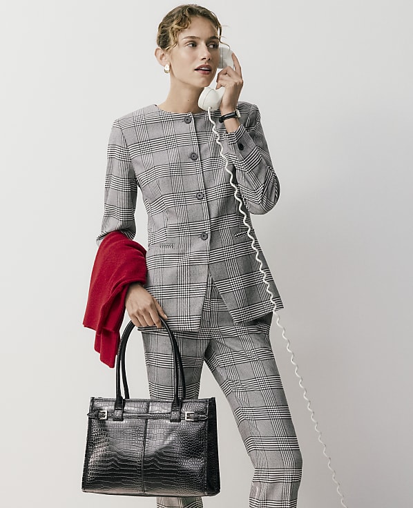 A woman in a grey plaid suit holds a white telephone and a black handbag against a plain white background.