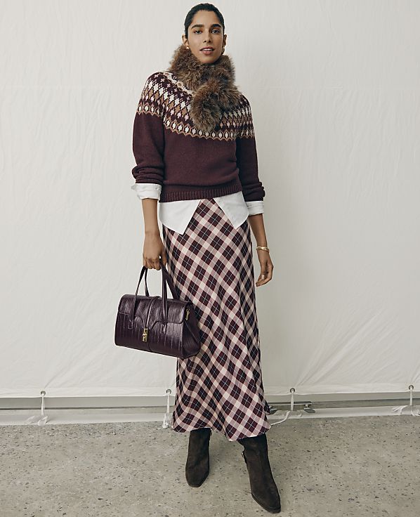 A woman wearing a patterned sweater, a plaid skirt, and a fur scarf stands holding a burgundy handbag against a plain white backdrop.