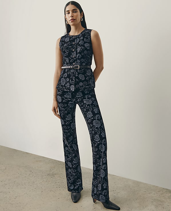 A woman wearing a dark floral print sleeveless top and matching trousers stands against a plain white wall and a light grey floor.