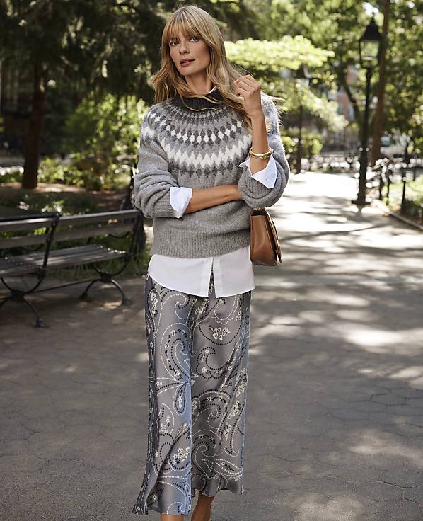 A woman wearing a grey patterned sweater and a paisley skirt stands on a paved path in a sun-dappled park with a wooden bench and lush greenery in the background.