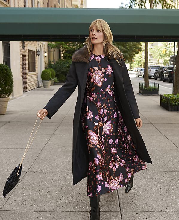 A woman in a floral dress and black coat stands on a city sidewalk with buildings and parked cars in the background.