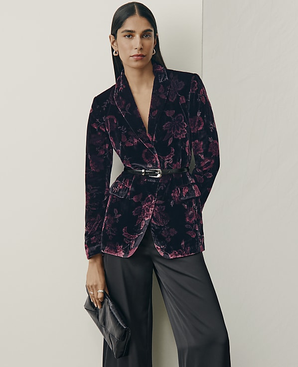 A woman wearing a floral purple velvet blazer and black trousers stands against a plain white wall.