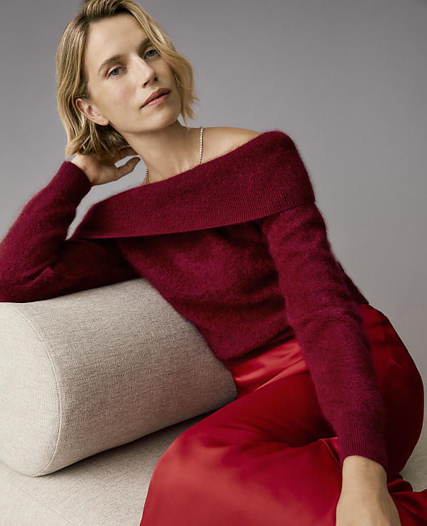 A woman wearing a burgundy off-the-shoulder sweater and red satin trousers leans against a beige bolster pillow against a plain grey background.