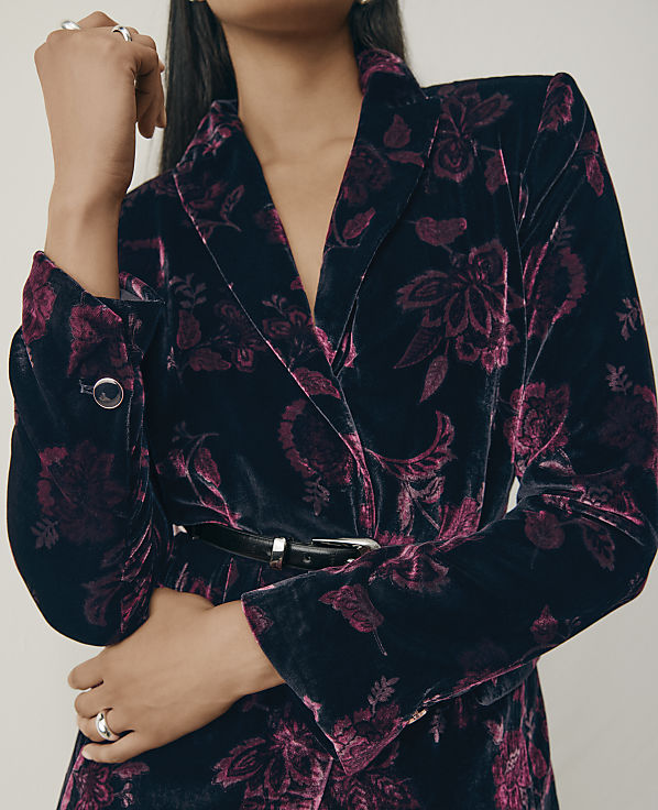 A person wearing a dark blue and purple floral velvet blazer and rings stands against a plain, light-colored background.