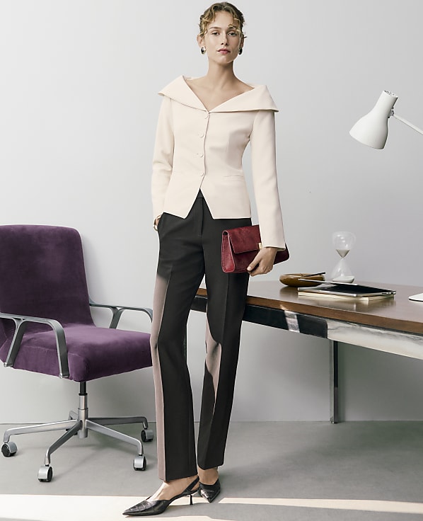 A woman in a pale pink blazer and black trousers stands holding a red clutch in an office setting with a purple chair and a desk.
