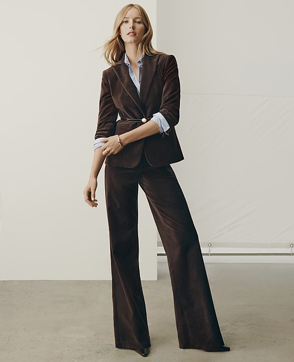 A woman wearing a brown velvet suit and a light blue shirt stands against a plain white studio background.