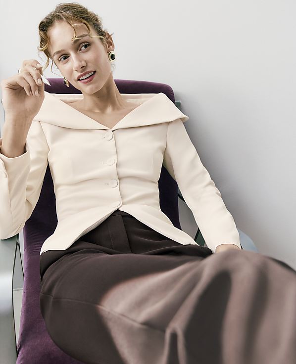 A woman wearing a cream-colored jacket and brown trousers sits in a purple chair against a plain white wall.