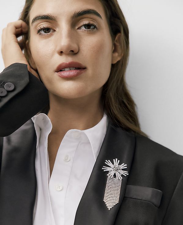 A woman wearing a black blazer with a sparkling brooch and a white shirt poses against a plain white background.