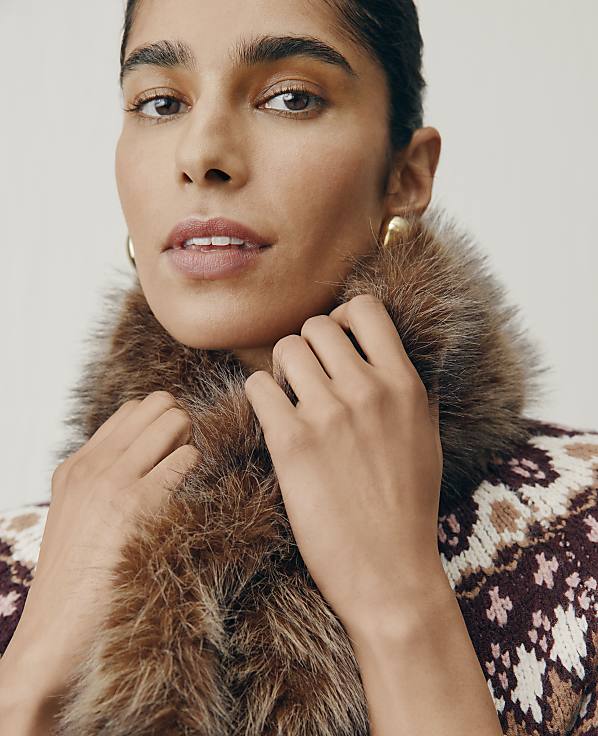 A woman wearing a brown faux-fur scarf and a patterned sweater poses against a plain, light-colored background.