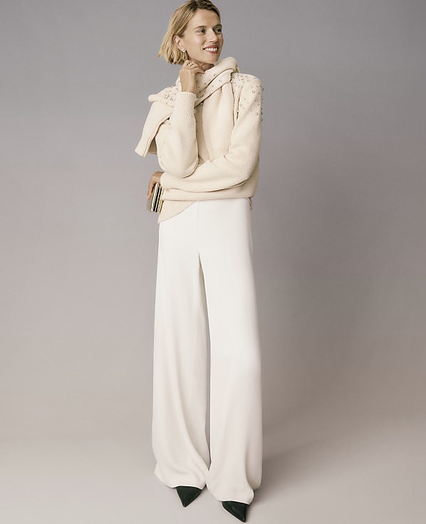 A woman wearing a cream-colored sweater and wide-leg white trousers stands against a plain grey background.