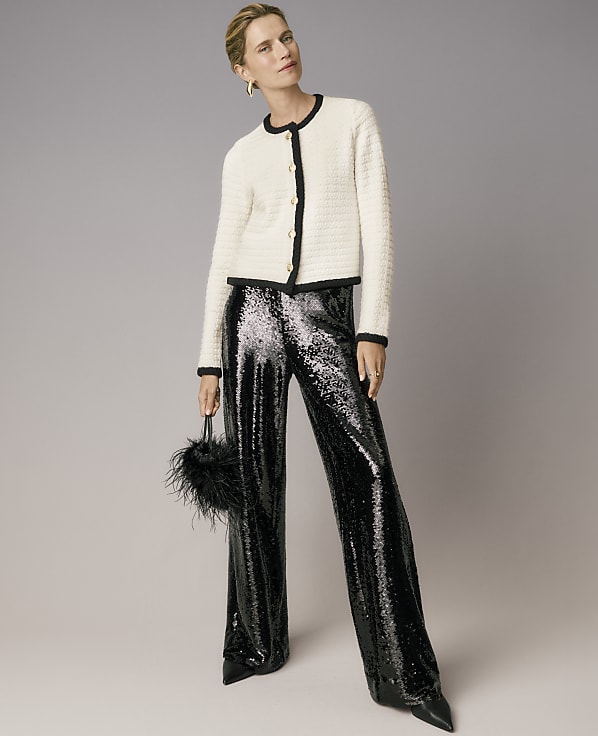 A woman wearing a white tweed jacket and shimmering black sequined trousers stands against a plain grey background.